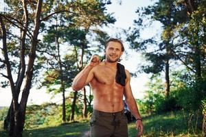 Close up portrait of woodsman with axe in hand. Handsome shirtless man with muscular body type is in the forest at daytime photo