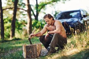 sostiene hacha de madera. un hombre guapo sin camisa con un tipo de cuerpo musculoso está en el bosque durante el día foto