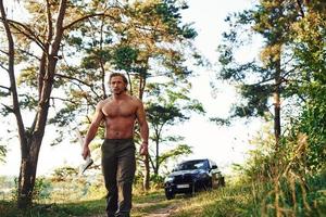 With axe. Handsome shirtless man with muscular body type is in the forest at daytime photo