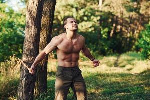 Doing workout. Handsome shirtless man with muscular body type is in the forest at daytime photo