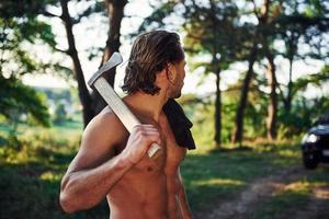 Close up portrait of woodsman with axe in hand. Handsome shirtless man with muscular body type is in the forest at daytime photo