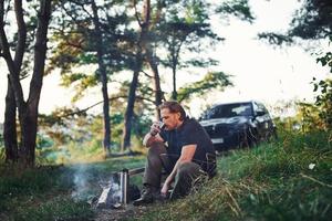 Enjoying fresh drink. Man in black shirt near the campfire in the forest at his weekend time photo