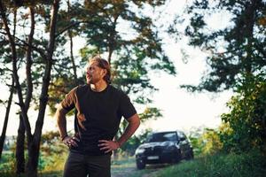 automóvil negro detrás. retrato del hombre que se para en el bosque y mira la naturaleza foto