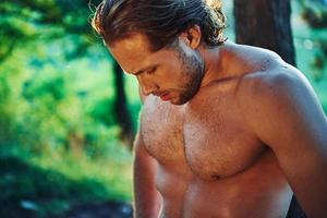 Serious and confident look. Handsome shirtless man with muscular body type is in the forest at daytime photo