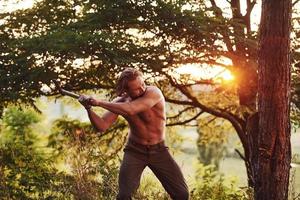 Process of cutting the tree by using the axe. Handsome shirtless man with muscular body type is in the forest at daytime photo