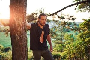 Leaning on the tree. Amazing sunlight. Handsome man with muscular body type is in the forest at daytime photo