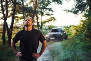 Black automobile behind. Portrait of man that stands in the forest and looks at nature photo