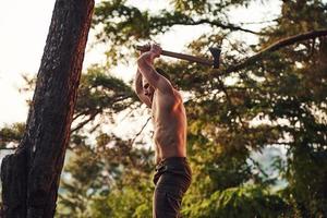 proceso de cortar el árbol usando el hacha. un hombre guapo sin camisa con un tipo de cuerpo musculoso está en el bosque durante el día foto