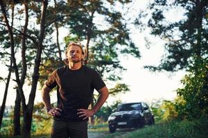 automóvil negro detrás. retrato del hombre que se para en el bosque y mira la naturaleza foto