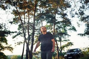 automóvil negro detrás. retrato del hombre que se para en el bosque y mira la naturaleza foto