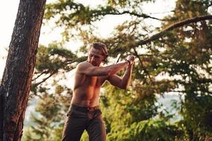 Process of cutting the tree by using the axe. Handsome shirtless man with muscular body type is in the forest at daytime photo