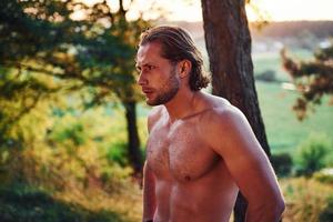 mirada seria y confiada. un hombre guapo sin camisa con un tipo de cuerpo musculoso está en el bosque durante el día foto