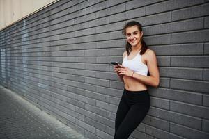 Positive girl smiling. Young sportive brunette with slim body shape against brick wall in the city at daytime photo