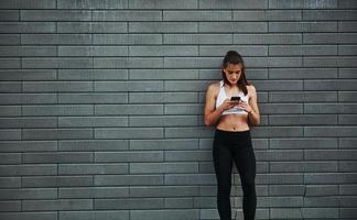 Using smartphone. Young sportive brunette with slim body shape against brick wall in the city at daytime photo