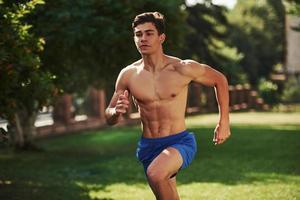 Going forward. Muscular shirtless man have fitness day and running in the park at daytime photo