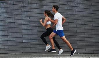 Healthy lifestyle. Man and woman have fitness day and running in the city at daytime near brick wall photo