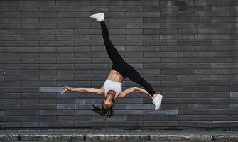 Doing crazy parkour and athletic stunts. Young sportive brunette with slim body shape against brick wall in the city at daytime photo