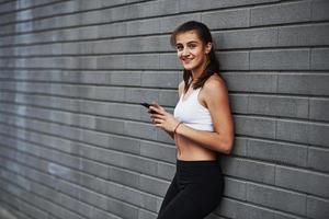 Cheerful mood. With smartphone in hands. Young sportive brunette with slim body shape against brick wall in the city at daytime photo