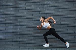 Running fast. Young sportive brunette with slim body shape against brick wall in the city at daytime photo