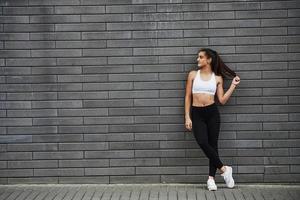 Young sportive brunette with slim body shape against brick wall in the city at daytime photo