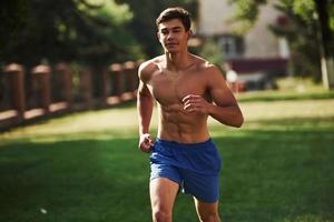 Going forward. Muscular shirtless man have fitness day and running in the park at daytime photo