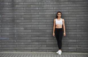 Young sportive brunette with slim body shape against brick wall in the city at daytime photo