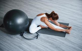 Sitting on roll mat. Young sporty woman in white clothes with slim body type in the healthy center doing exercises. Black fitness ball photo