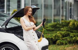 Leans on the front of the automobile. Woman on the electric cars charge station at daytime. Brand new vehicle photo