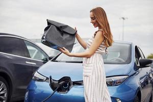 sostiene una bolsa negra. mujer en la estación de carga de coches eléctricos durante el día. vehículo nuevo foto