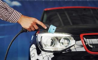 Man stands against an electric car and holds the charging cable photo