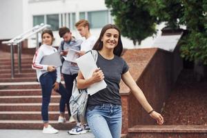 Positive girl in front of friends. Group of young students in casual clothes near university at daytime photo