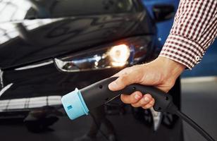 Man stands against an electric car and holds the charging cable photo