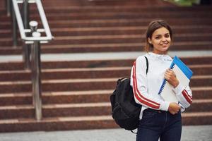 Positive female student standing outdoors with documents at daytime photo