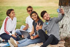 sentado cerca del árbol y haciendo selfie. grupo de jóvenes estudiantes con ropa informal sobre hierba verde durante el día foto