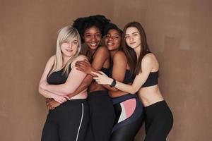 abrazos y sonrisas. grupo de mujeres multiétnicas de pie en el estudio con fondo marrón foto