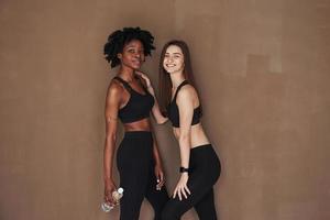 Modern sportive people. Two multi ethnic female friends stands in the studio with brown background photo
