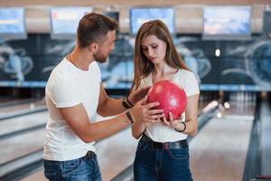 Man teaching girl how to holds ball and play bowling in the club photo