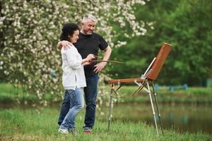 Standing near the lake. Mature couple have leisure days and working on the paint together in the park photo