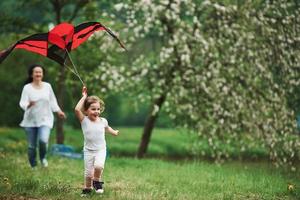 Summertime happiness. Positive female child and grandmother running with red and black colored kite in hands outdoors photo