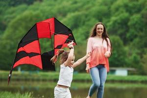Two females. Positive child and her mother running with red and black colored kite in hands outdoors photo