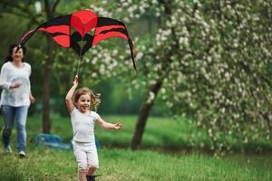 Woman is behind. Positive female child and grandmother running with red and black colored kite in hands outdoors photo