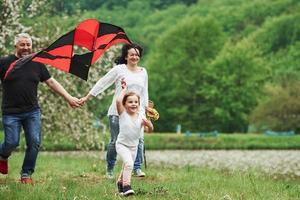 Awesome feeling. Positive female child with grandmother and grandfather running with red and black colored kite in hands outdoors photo