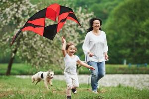 Two people and dog. Positive female child and grandmother running with red and black colored kite in hands outdoors photo