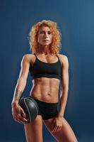 Stands with black soccer ball. Redhead female bodybuilder is in the studio on blue background photo