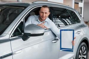 Showing by forefinger. Manager sitting in modern white car with paper and documents in hands photo