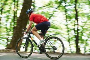 Sportive clothes. Cyclist on a bike is on the asphalt road in the forest at sunny day photo