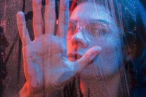 tocando el vaso con la mano. foto de estudio en estudio oscuro con luz de neón. retrato de hombre serio detrás del vidrio mojado