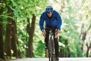 In blue jacket. Cyclist on a bike is on the asphalt road in the forest at sunny day photo