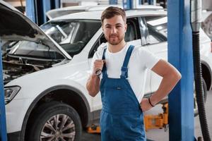 de pie con una llave en la mano. empleado en el uniforme de color azul trabaja en el salón del automóvil foto