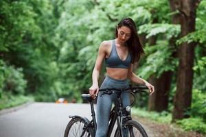 Calm and quiet. Female cyclist standing with bike on asphalt road in the forest at daytime photo